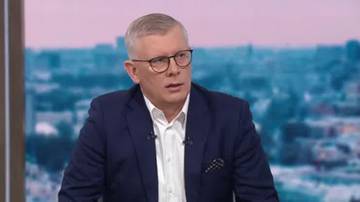 Mężczyzna w okularach i białej koszuli, ubrany w granatową marynarkę, z zapiętą krawatką, siedzi przed ekranem z rozmytym obrazem miasta.