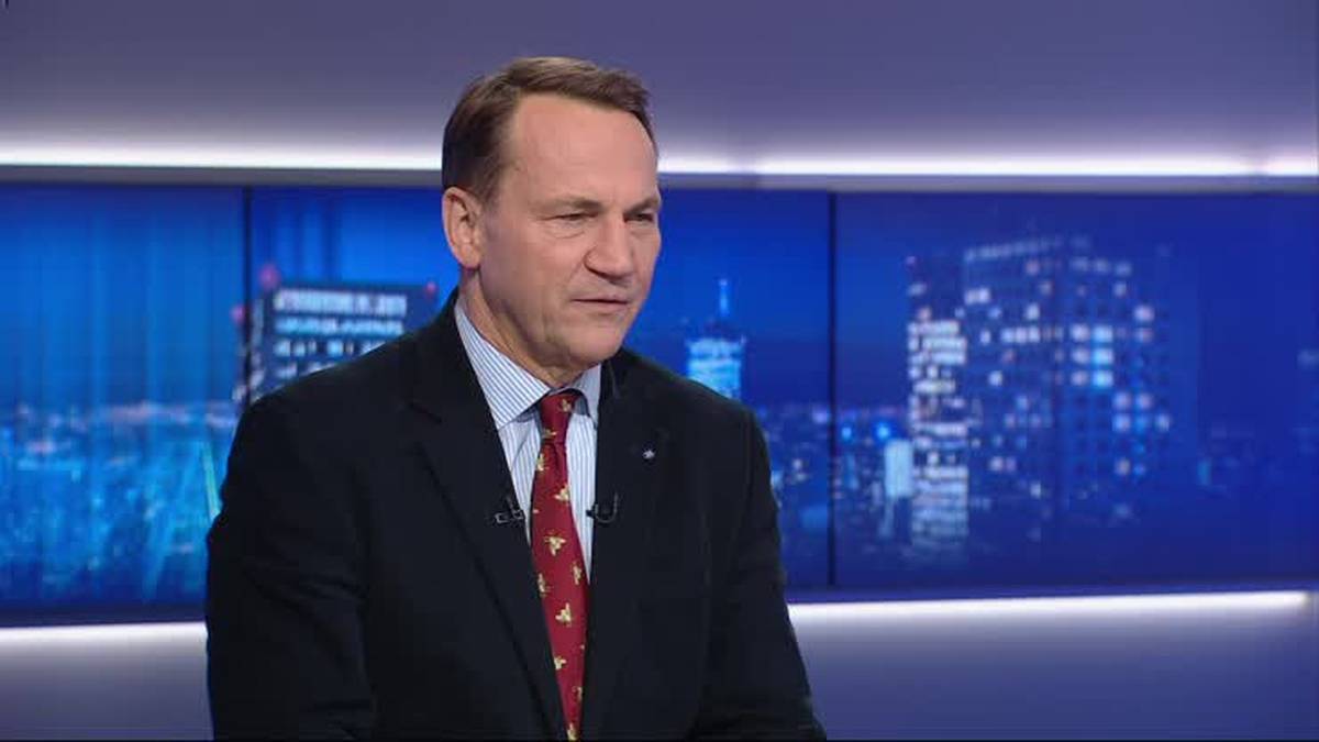 Radosław Sikorski w garniturze przed panelem świetlnym, z nocnym miastem w tle.