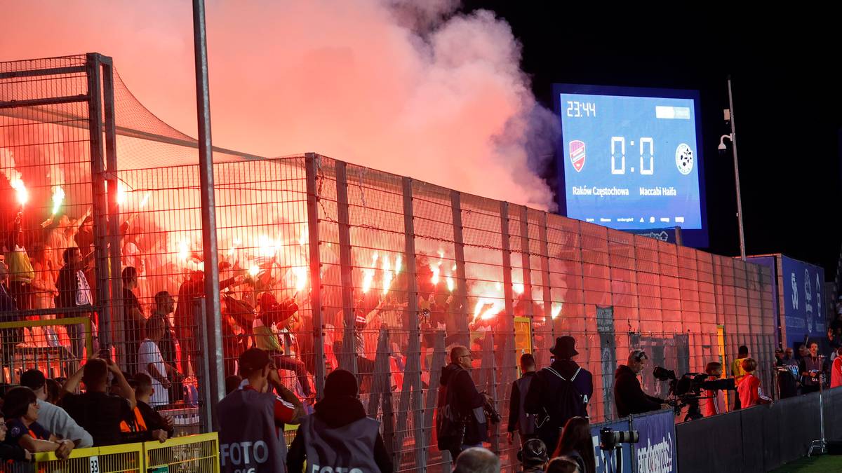 Widzowie na nocnym meczu piłki nożnej odpalają race, tworząc czerwone zadymienie. Na wielkim ekranie widoczny jest wynik 0:0.
