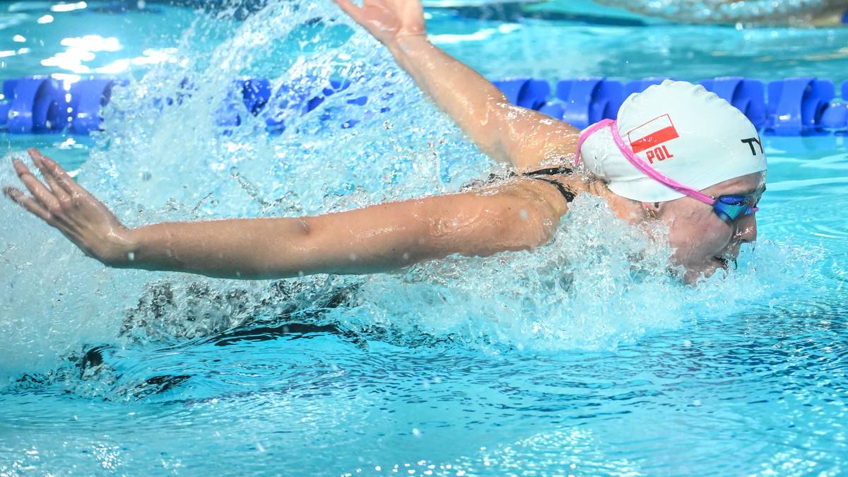 Pływaczka w białym czepku z biało-czerwoną flagą Polski wykonuje styl motylkowy w basenie.
