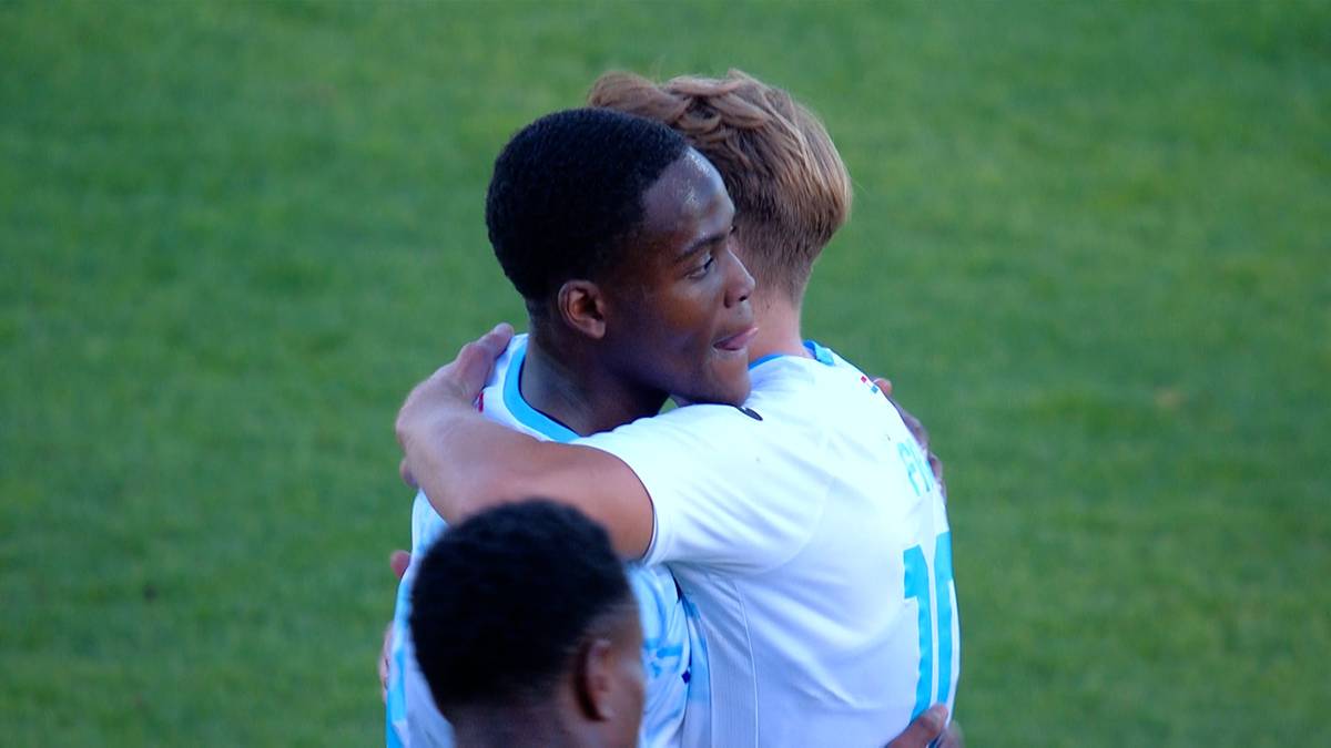 Dw&oacute;ch piłkarzy w białych strojach obejmuje się na zielonym boisku piłkarskim.