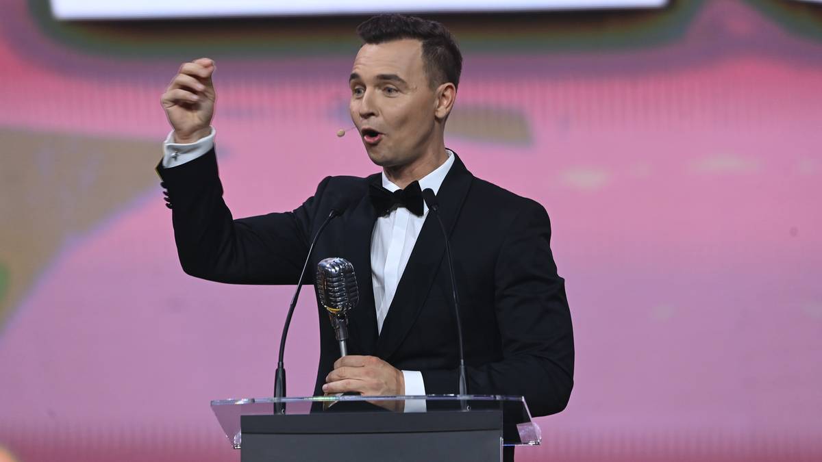 Jerzy Mielewski w czarnym garniturze i muszce przemawia przy mównicy z mikrofonem.