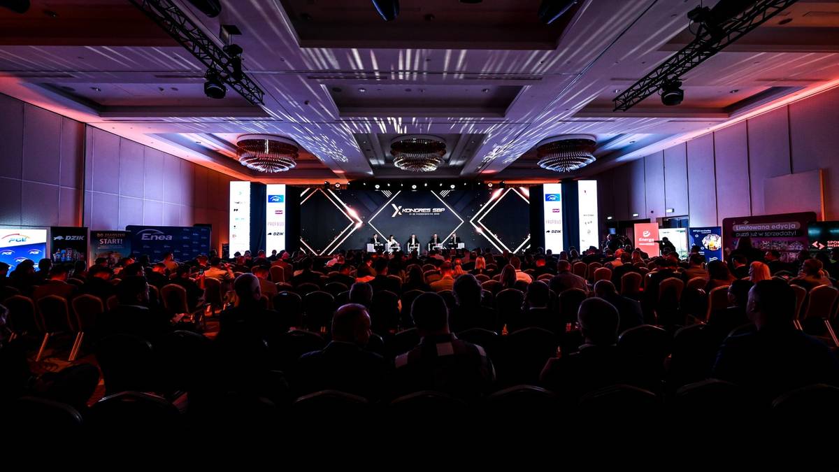 Widok z tyłu sali konferencyjnej na scenę z ekranem i oświetleniem, wypełnioną liczną publicznością.