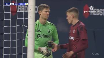 Bramkarz w zielonej koszulce i zawodnik w czerwonej koszulce rozmawiają obok bramki podczas meczu piłki nożnej.