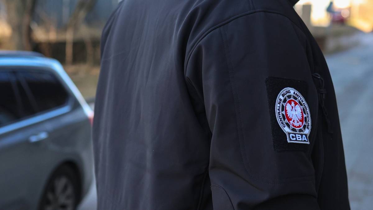 Fragment kurtki z naszywką Centralnego Biura Antykorupcyjnego (CBA) w tle samochód