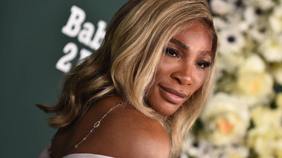 Serena Williams, uśmiechnięta, patrzy w stronę obiektywu. Ma jasne blond włosy i nosi jasnoróżową sukienkę. W tle widoczne są białe kwiaty i zielona ściana z napisem "Bal 27".