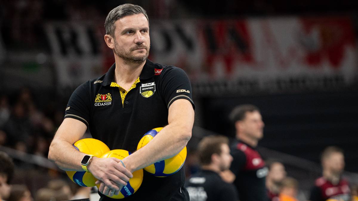 Trener siatkówki w czarnej koszulce z żółtymi elementami, trzymający w rękach piłki siatkowe.