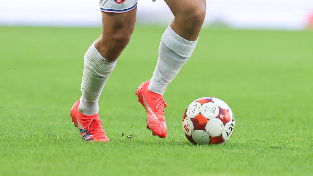 Fragment ciała piłkarza, widoczne nogi w białych skarpetach i pomarańczowych butach, z piłką nożną na zielonej trawie.