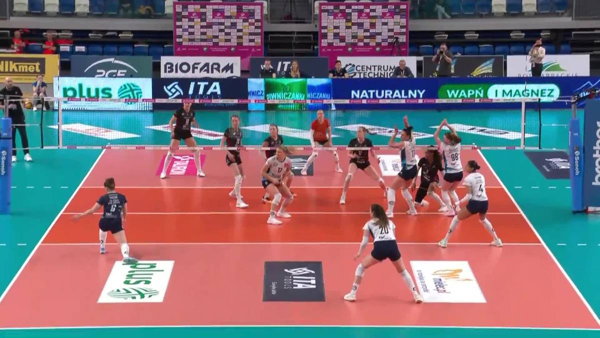 Zawodniczki grające w siatk&oacute;wkę podczas meczu w hali sportowej. Widoczne są dwie drużyny w kontrastujących strojach, rozgrywające piłkę nad siatką. W tle znajdują się trybuny z publicznością oraz banery reklamowe.