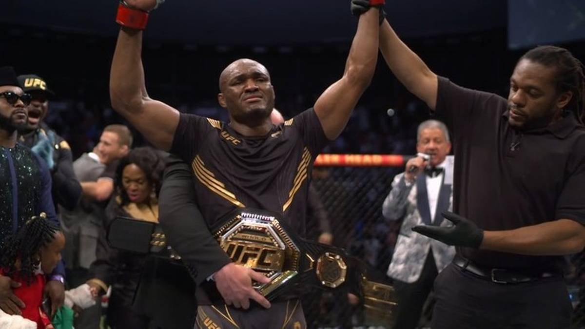 Kamaru Usman, były mistrz UFC, podnosi ręce w geście triumfu z pasem mistrzowskim na biodrach. Po jego prawej stronie stoi mężczyzna w czarnym ubraniu, również unoszący rękę.