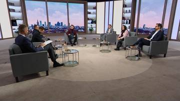 Siedem os&oacute;b w okręgu, dyskutujących w studio telewizyjnym z widokiem na miasto w tle.