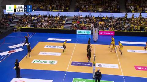 Sześciu siatkarzy w czarnych i ż&oacute;łtych strojach gra mecz siatk&oacute;wki na niebieskim boisku. Widownia wypełnia trybuny w tle.