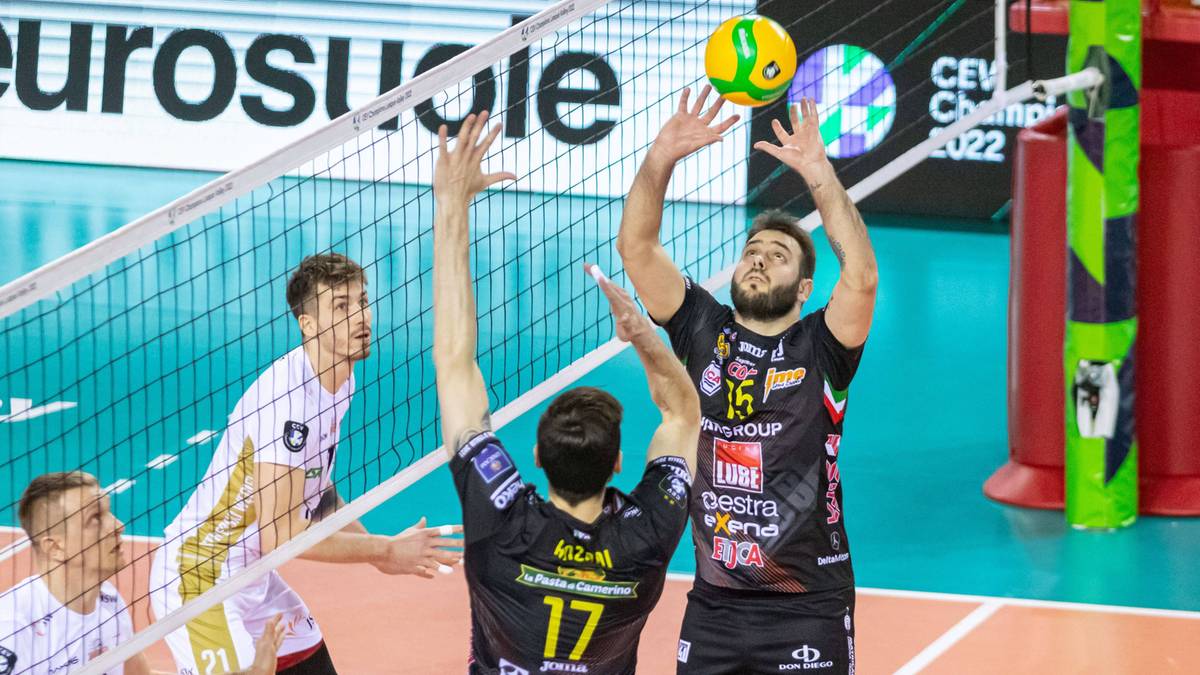 Siatkarz w skoku atakuje piłkę nad siatką. Na drugim planie widoczni są inni zawodnicy oraz siatka.