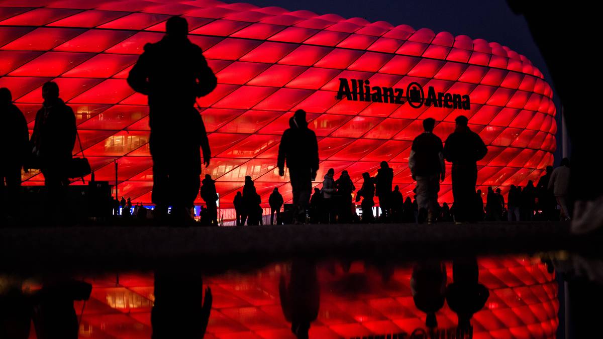 Widok na Allianz Arenę nocą, podświetloną na czerwono. Widać sylwetki ludzi na pierwszym planie, a ich odbicie w kałuży lub mokrej nawierzchni.