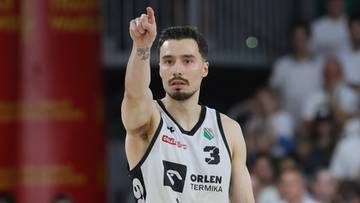 Koszykarz w stroju sportowym z numerem 3 na koszulce, trzymający piłkę i wskazujący palcem w g&oacute;rę.