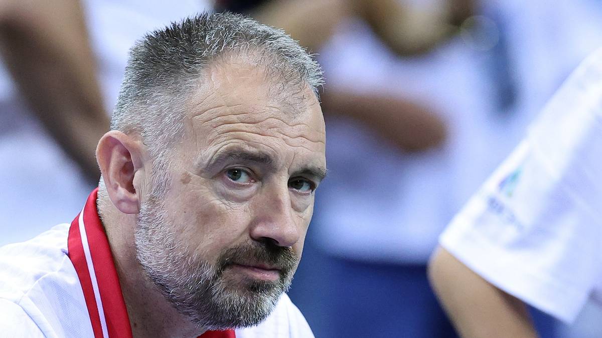 Portret starszego mężczyzny z kr&oacute;tkimi siwymi włosami i brodą, w białej koszulce i z czerwoną wstążką na szyi.