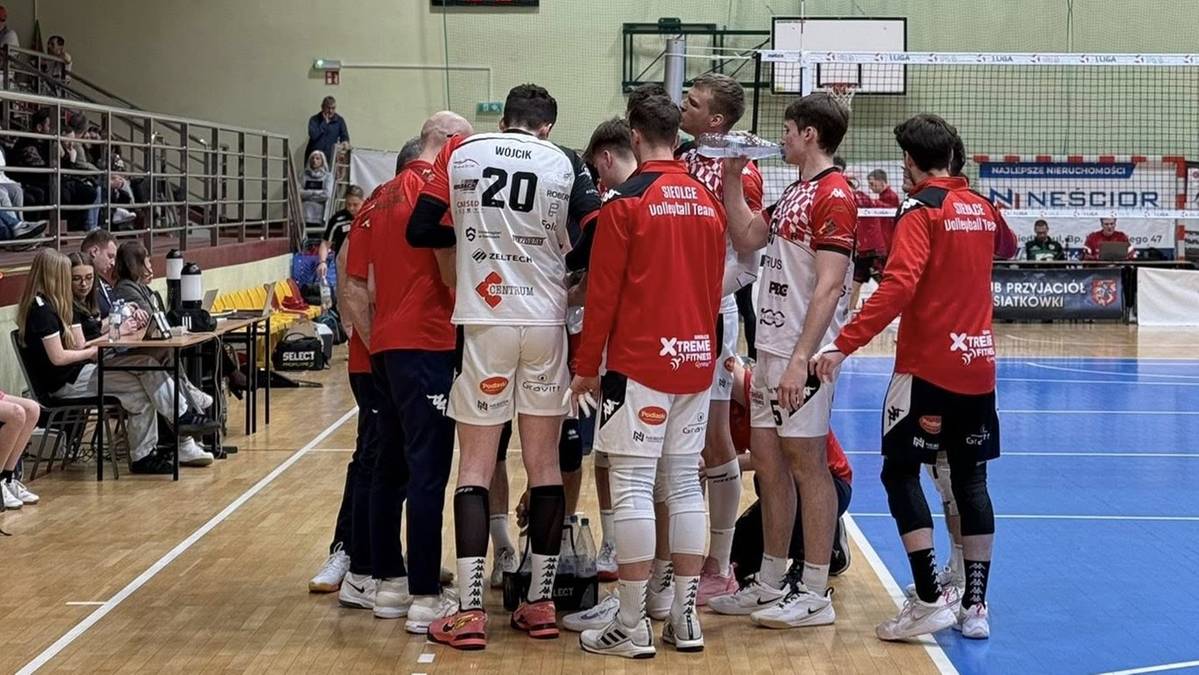 Drużyna siatkarzy w strojach sportowych stoi w k&oacute;łku na boisku, koncentrując się przed meczem lub po przerwie.