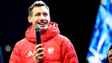 Kacper Tomasiak w kurtce z godłem Polski, trzymający mikrofon i uśmiechający się.