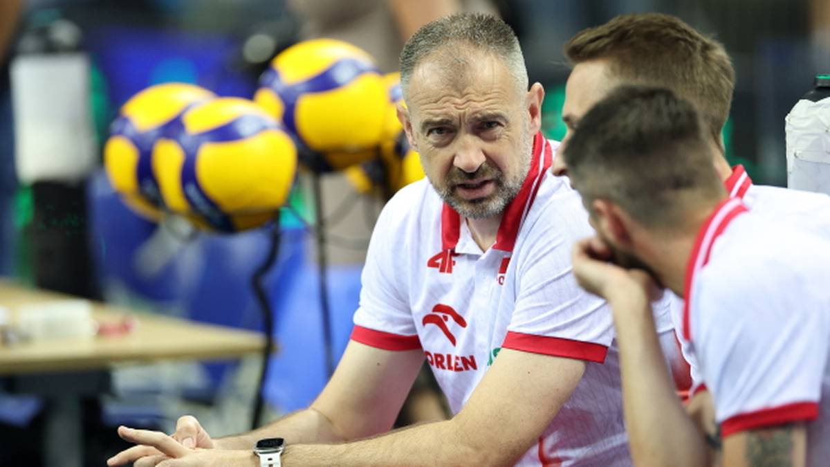 Trener siatkówki w białej koszulce z czerwonymi elementami rozmawia z dwoma zawodnikami.