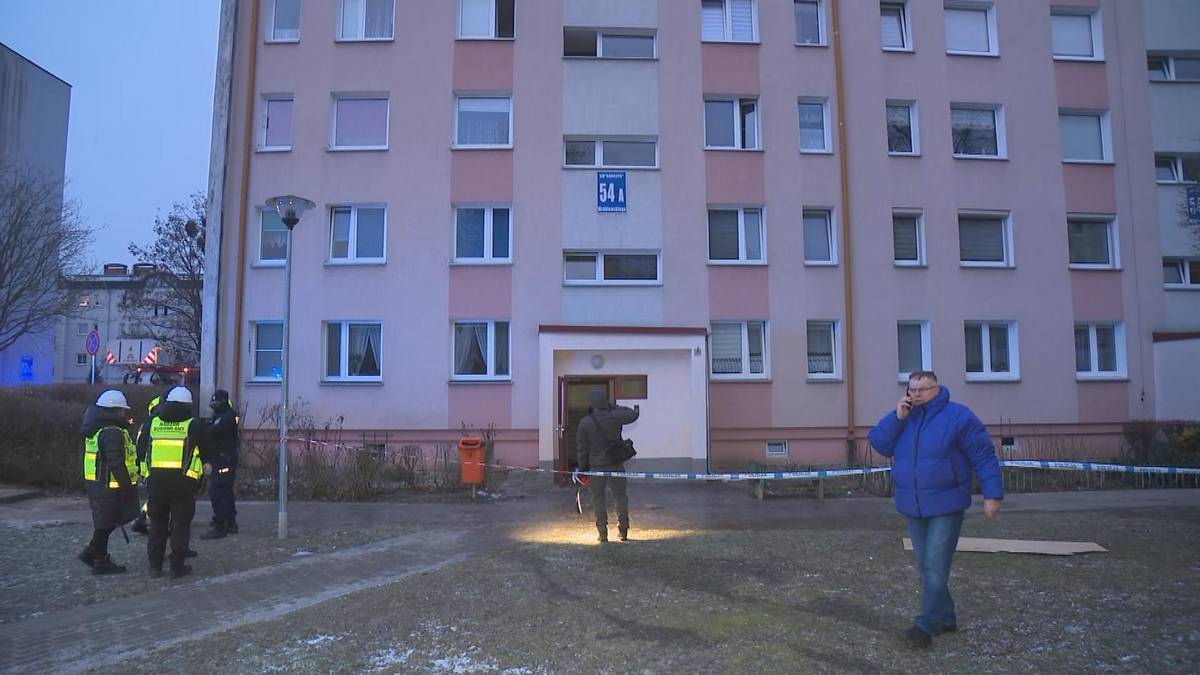 Funkcjonariusze policji i straży pożarnej przy wielopiętrowym bloku mieszkalnym, teren zabezpieczony taśmą.