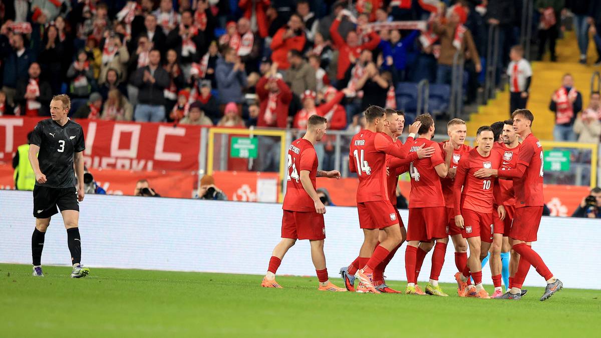 Piłkarze w czerwonych strojach świętują zdobycie gola, na pierwszym planie jeden piłkarz w czarnym stroju opiera się o nogę.