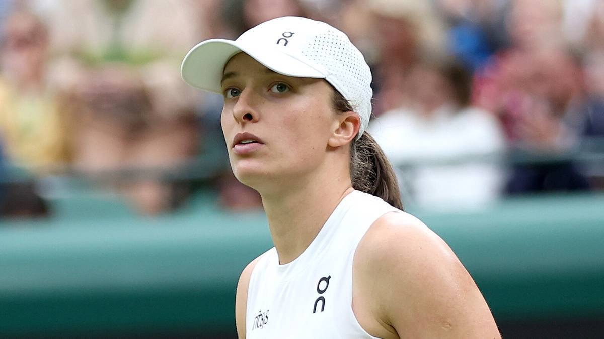 Zbliżenie na twarz Igi Świątek, polskiej tenisistki, w białej czapce z daszkiem i białej koszulce sportowej. Jest skupiona i patrzy w bok. W tle rozmyty tłum widzów.