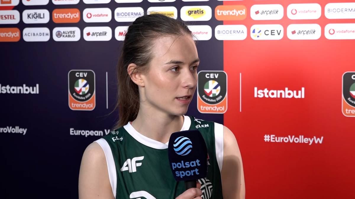 Młoda kobieta w zielonym stroju sportowym z mikrofonem w dłoni, udziela wywiadu na tle baneru z logo turnieju siatk&oacute;wki.