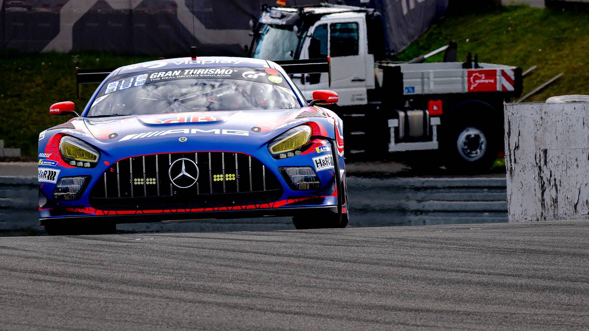 Niebieski samoch&oacute;d wyścigowy z czerwono-ż&oacute;łtymi akcentami na torze wyścigowym, z ciężar&oacute;wką na dalszym planie.