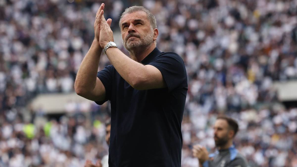 Trener piłkarski w czarnej koszulce z krótkim rękawem klaszcze w dłonie, stojąc na tle stadionu wypełnionego kibicami.