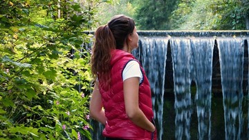 Kobieta w czerwonej kamizelce patrzy na spadającą wodę z tamy w otoczeniu zielonej roślinności.