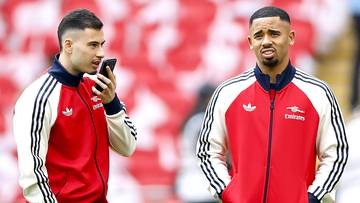 Dw&oacute;ch piłkarzy w czerwonych bluzach sportowych, jeden rozmawia przez telefon, drugi spogląda przed siebie. W tle trybuny stadionu.