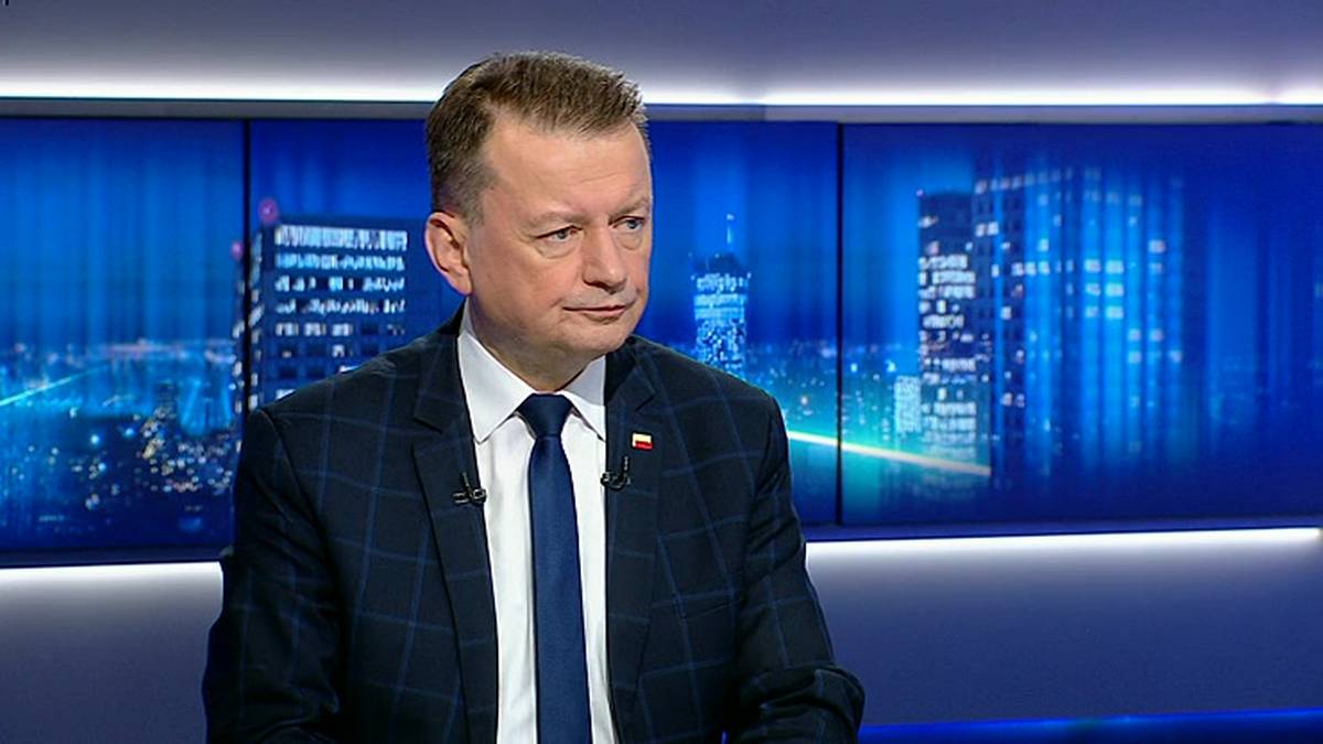 Mężczyzna w garniturze i krawacie siedzi przed niebieskim tłem z widokiem na nocne miasto.