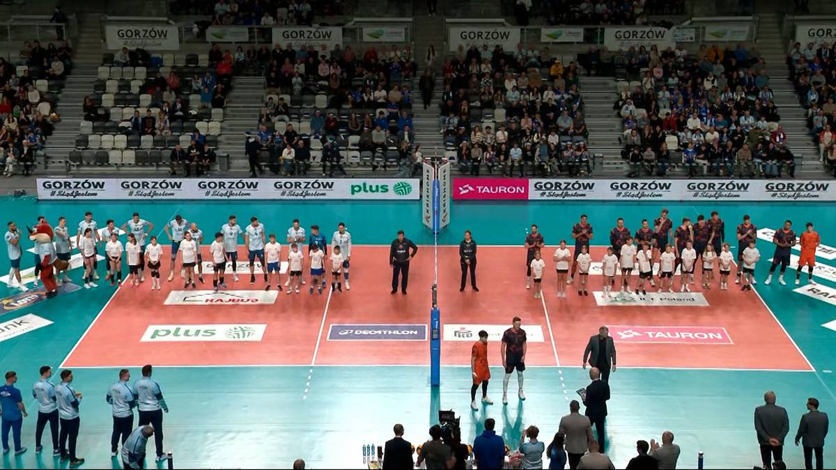 Dwie drużyny siatkarzy stoją naprzeciwko siebie na boisku, otoczone przez trybuny wypełnione widzami.
