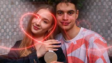 Młoda para z medalem. Kobieta z medalem w dłoni opiera się o ramieniem mężczyzny. Nad nimi widnieje czerwony zarys serca.