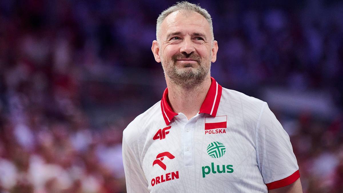 Średniozbliżenie na mężczyznę w białej koszulce z czerwonymi elementami i logo reprezentacji Polski. Mężczyzna ma krótkie, siwe włosy, zarost i uśmiecha się.