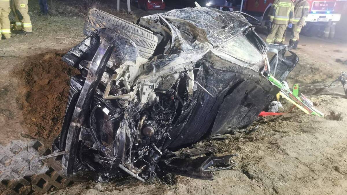 Zniszczone BMW leżące na boku po wypadku drogowym, z widocznymi służbami ratowniczymi w tle.
