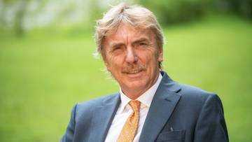 Mężczyzna w średnim wieku o blond włosach i wąsach, ubrany w ciemny garnitur i pomarańczowy krawat, uśmiecha się do aparatu.