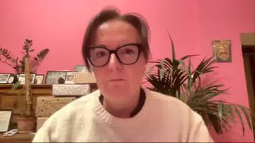 Joanna Sadzik w okularach na tle różowej ściany z roślinami.