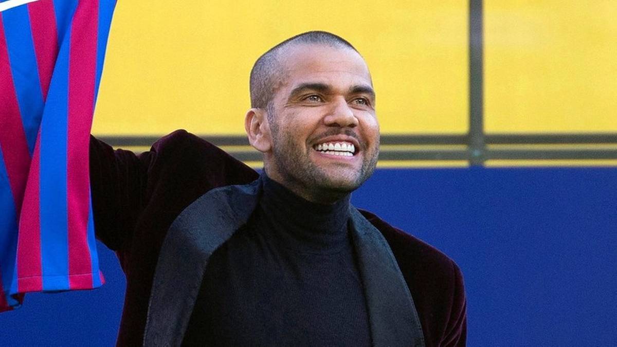 Uśmiechnięty mężczyzna w czarnym golfie i marynarkowym szalu, z fragmentem flagi w niebiesko-r&oacute;żowe pasy.