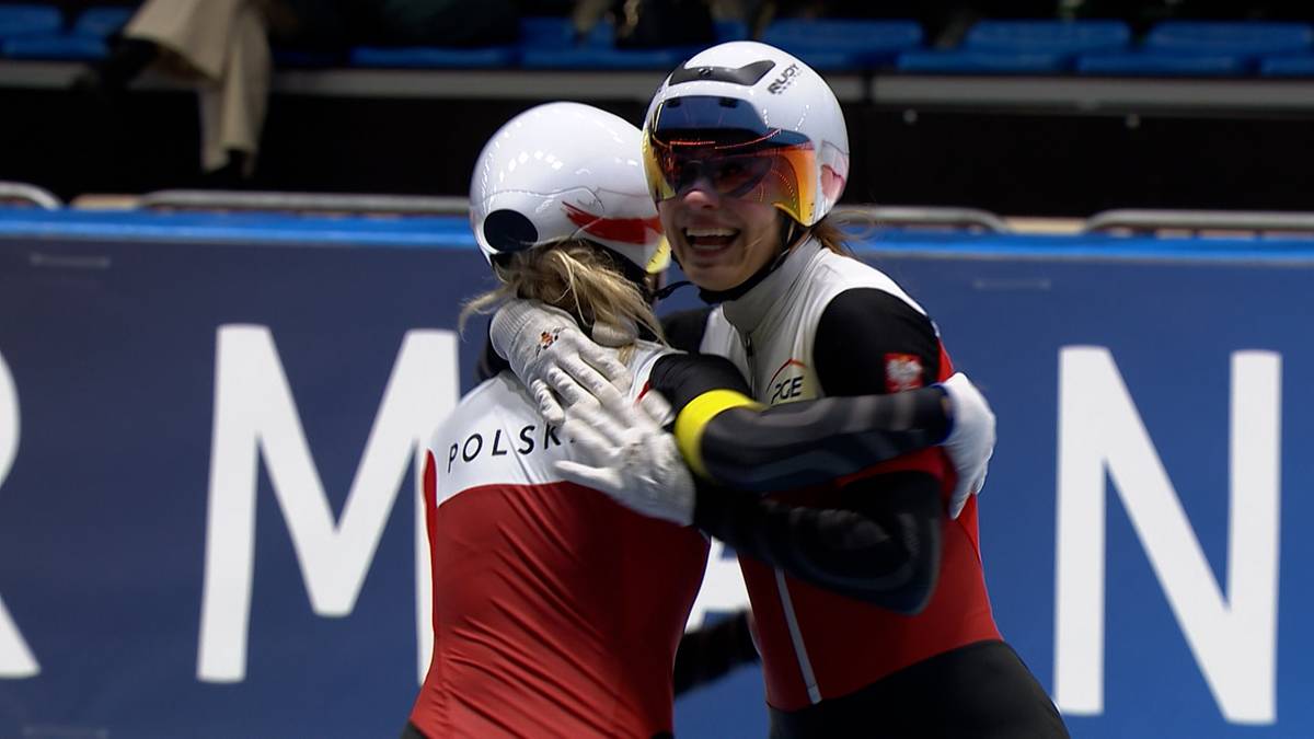 Dwie panczenistki w kaskach i strojach sportowych przytulają się po zawodach, jedna z nich ma na stroju napis "POLSKA".
