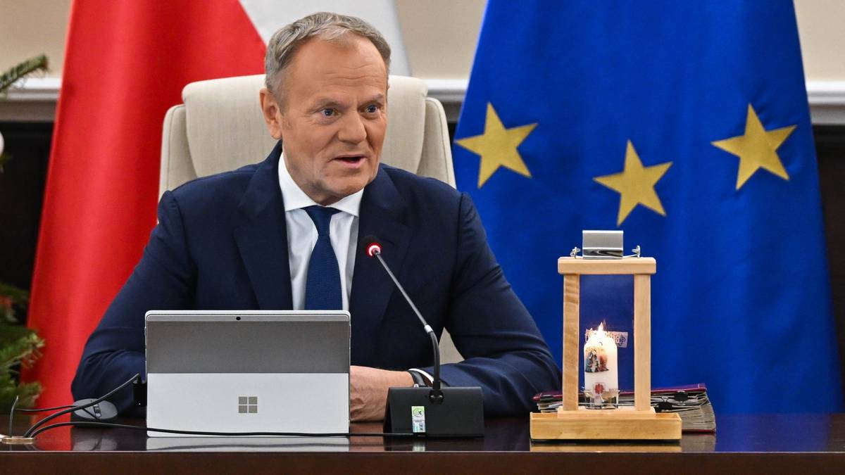 Premier Donald Tusk siedzi przy biurku, na kt&oacute;rym znajduje się laptop i świeca w lampionie. W tle widoczne są flagi Polski i Unii Europejskiej.