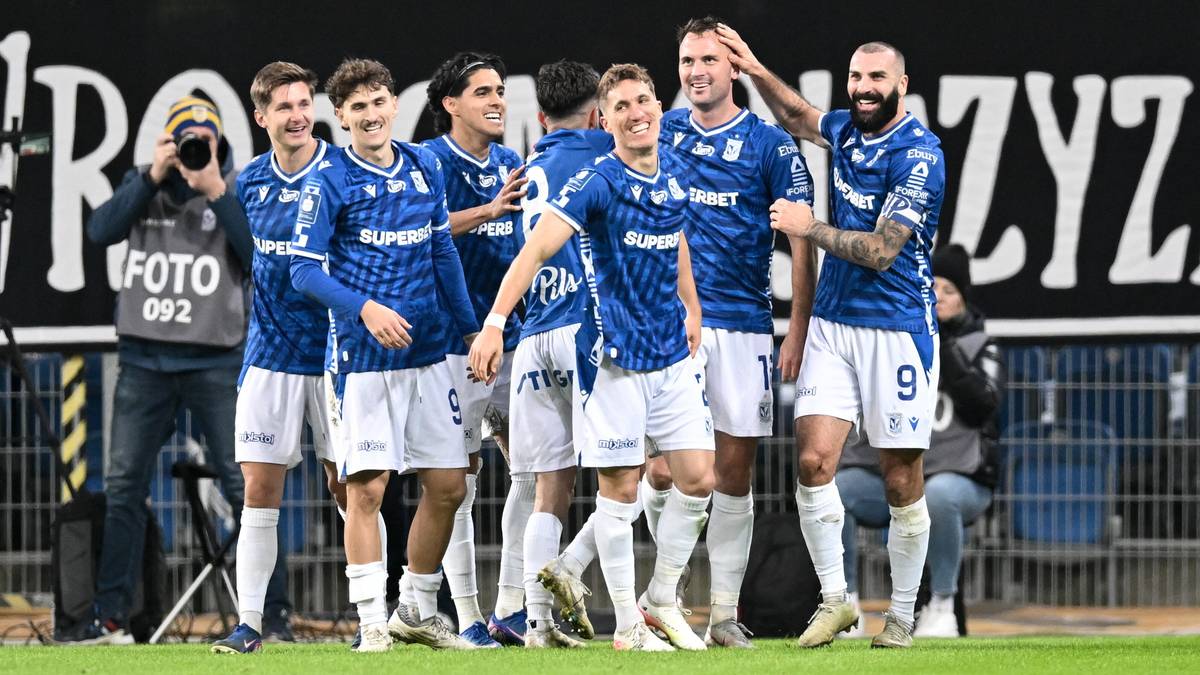 Grupa piłkarzy w niebiesko-białych strojach sportowych, stojących obok siebie na boisku, pozuje do zdjęcia. Jeden z zawodnik&oacute;w z tyłu trzyma aparat fotograficzny. Wszyscy wyglądają na szczęśliwych.