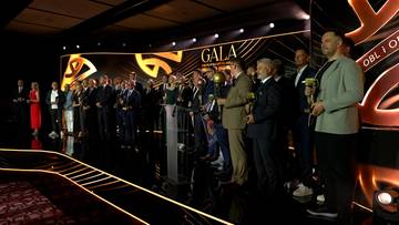 Grupa os&oacute;b w eleganckich strojach stoi na scenie podczas gali, część z nich trzyma nagrody. W tle widoczny napis "GALA" i dekoracje.