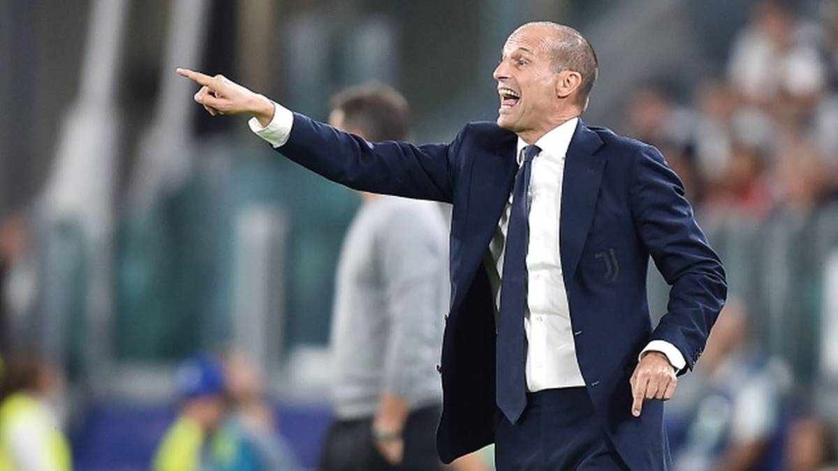 Massimiliano Allegri, trener piłkarski, ubrany w ciemny garnitur i białą koszulę z krawatem, wskazuje palcem w lewo i krzyczy, stojąc na boisku sportowym podczas meczu.