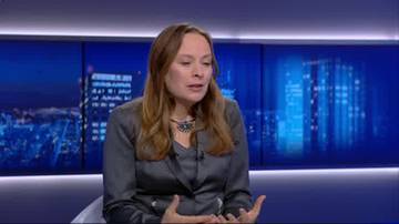 Kobieta z długimi, jasnobrązowymi włosami, ubrana w ciemnoszarą marynarkę, siedzi i m&oacute;wi. W tle widoczne są niebieskie ekrany z widokiem na nocną panoramę miasta.