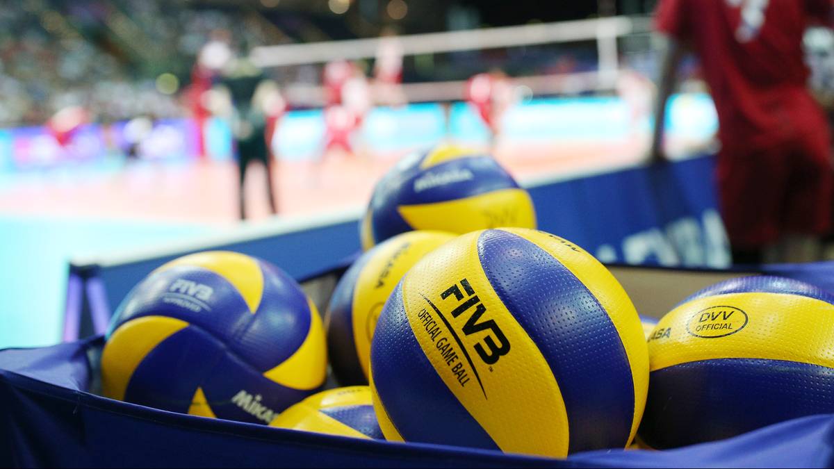 Piłki do siatkówki w niebiesko-żółte pasy leżą obok siebie w niebieskim pojemniku. W tle widać boisko do siatkówki z zawodnikami.