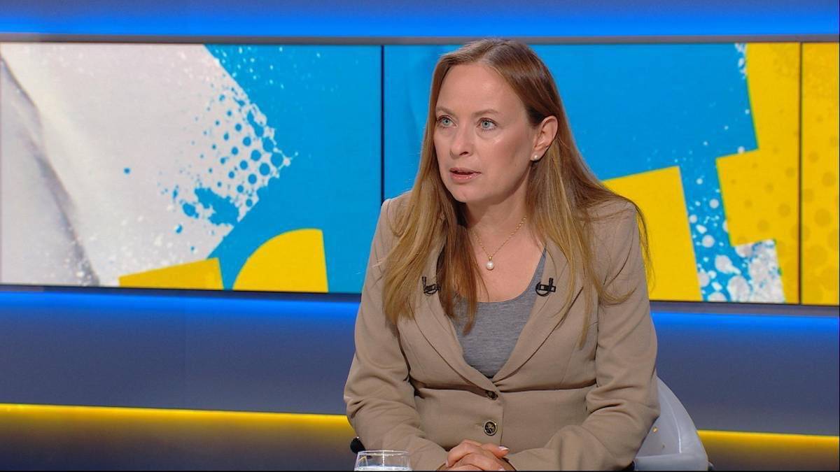 Kobieta w jasnobrązowym żakiecie siedzi przed kolorowym tłem.