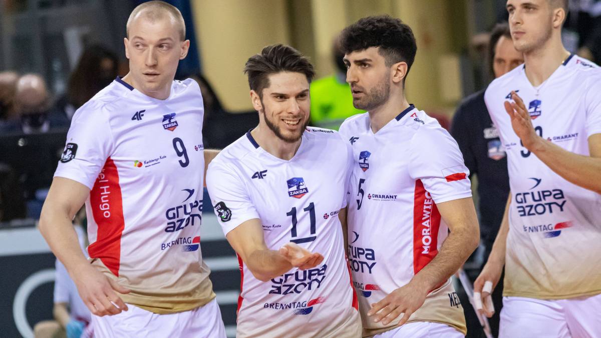 Siatkarze w białych koszulkach z numerami na plecach i czerwonymi elementami na rękawach, rozmawiający ze sobą podczas meczu.