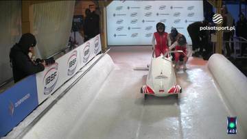 Dwie kobiety w kombinezonach narciarskich siedzą w dwuosobowej łodzi bobslejowej na torze lodowym, przygotowując się do startu. Jedna z nich, w czerwono-białym stroju, siedzi z przodu i trzyma kierownicę. Druga, w czarnym stroju, siedzi z tyłu. Na torze widać białe bandy z logotypami, a w tle kibiców i budynki.