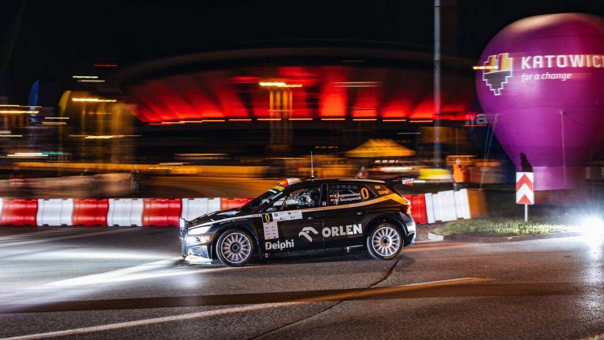 Samoch&oacute;d rajdowy ORLEN na trasie nocnego rajdu, z rozmytym tłem budynk&oacute;w i oświetlenia, sugerującym ruch i prędkość.
